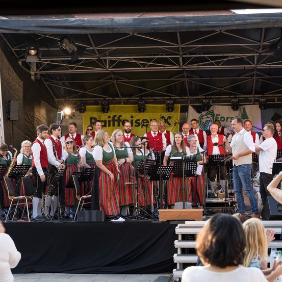 Ein Chor in traditionellen Kostümen steht auf der Bühne und führt ein Musikstück mit Musikinstrumenten auf, während ein Banner im Hintergrund zu sehen ist.