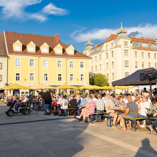 Eine große Gruppe von Menschen versammelt sich in einem Außencafé. Sie sitzen an Tischen unter gelben Sonnenschirmen. Eine Person im Rollstuhl ist ebenfalls anwesend. Im Hintergrund befinden sich Gebäude mit braunen Dächern und vielen Fenstern.