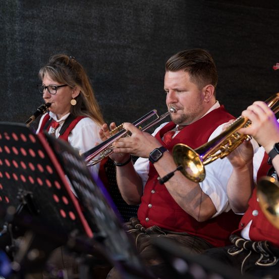 Eine Gruppe von Musikern spielt Instrumente. Eine Frau spielt die Klarinette, während zwei Männer Trompeten spielen. Sie tragen alle rote Westen und sitzen.