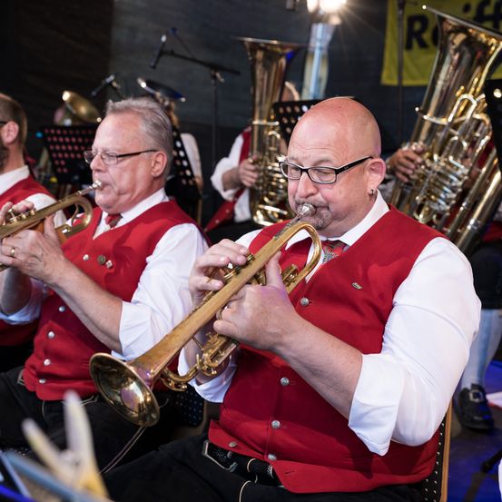 Eine Gruppe von Männern in roten Westen und weißen Langarmhemden spielt Blasinstrumente in einer Band. Ein Mann spielt die Trompete, während andere Posaunen und Tuben spielen.