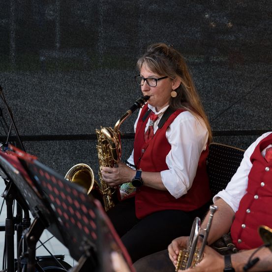 Eine Frau in einem roten Weste und einer Brille spielt Saxophon, während ein Mann in einem roten Weste die Trompete spielt. Sie sitzen beide vor einem Mikrofon.