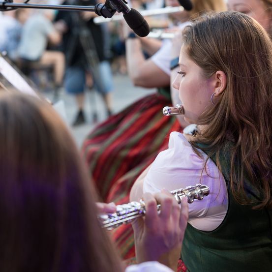 Eine junge Frau spielt eine Flöte bei einer Aufführung im Freien. Sie ist von anderen Musikern umgeben, und das Publikum im Hintergrund ist unscharf.