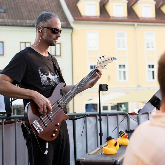 Ein Mann mit Sonnenbrille und Kopfhörern spielt eine Bassgitarre im Freien. Hinter ihm stehen Gummienten auf einem Tisch.