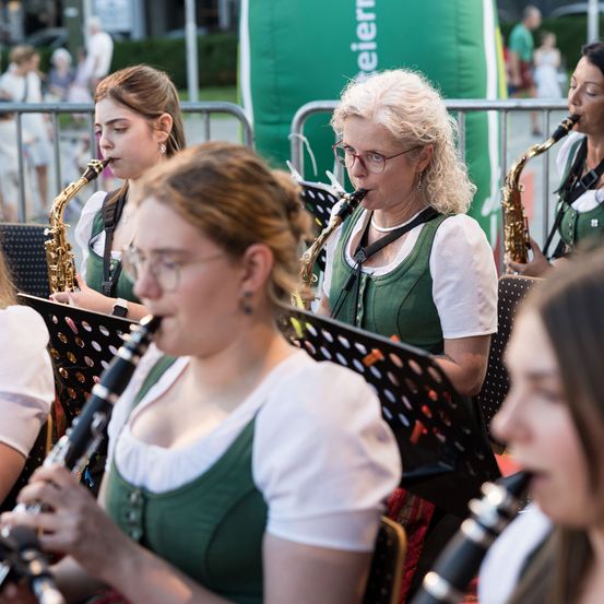 Eine Gruppe von Frauen in traditioneller grüner Kleidung spielt bei einer Veranstaltung im Freien. Sie spielen Musikinstrumente, darunter Saxophone und Klarinetten.