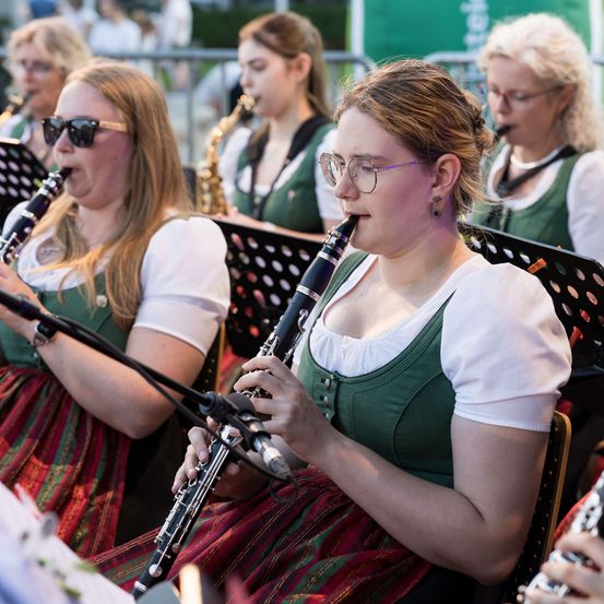 Mehrere Frauen in traditioneller Kleidung spielen Musikinstrumente, darunter Klarinetten, im Freien.