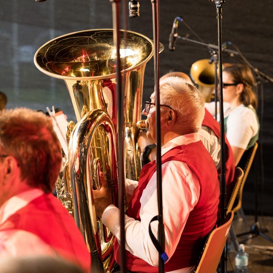 Ein Mann in einem roten Westchen spielt eine Messingtuba in einem Blasorchester während einer Aufführung. Eine Frau mit Brille sitzt hinter ihm, und ein Mikrofon steht auf einem Stativ.
