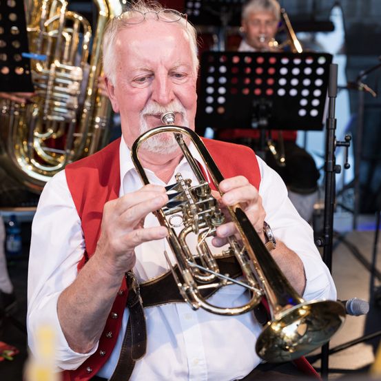 Ein älterer Mann spielt eine Trompete während einer musikalischen Aufführung. Er trägt ein rotes Weste und ein weißes Hemd, mit einem Mikrofon in der Nähe. Im Hintergrund spielt ein anderer Musiker ein Posaune.