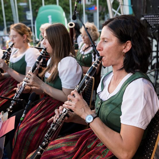 Eine Frau in einem grünen Kleid spielt Klarinette in einer Band mit anderen Frauen. Die Band spielt draußen.