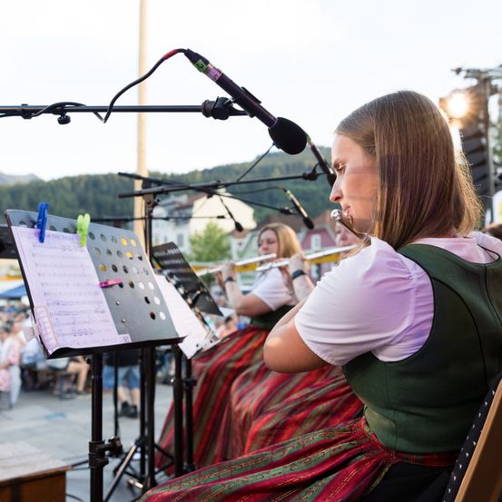 Ein Mädchen in traditioneller Kleidung spielt die Flöte bei einem Outdoor-Musikereignis. Ein Notenständer und ein Mikrofon sind in der Nähe, mit weiteren Musikern und einem Publikum im Hintergrund.