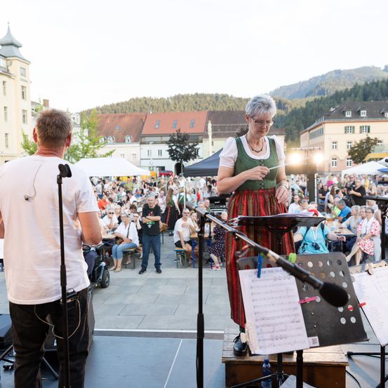 Ein Dirigent und Musiker auf einer Bühne mit Notenständern vor einem Publikum in einer Freiluftveranstaltung. Dahinter befinden sich Gebäude und eine malerische Kulisse.