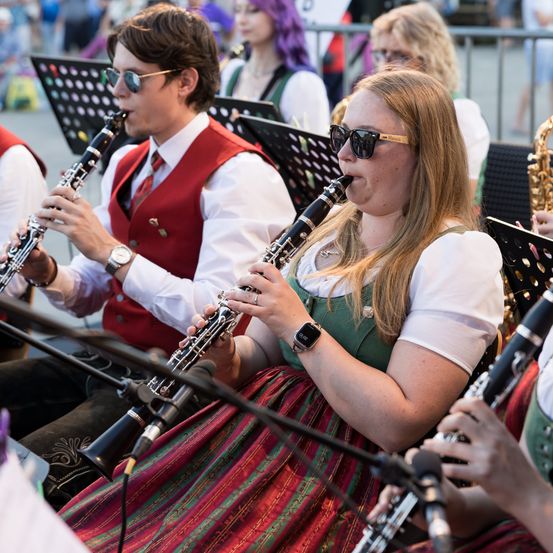 Eine Gruppe von Musikern spielt Klarinetten bei einer Open-Air-Veranstaltung, eine Frau in traditioneller Kleidung im Vordergrund.