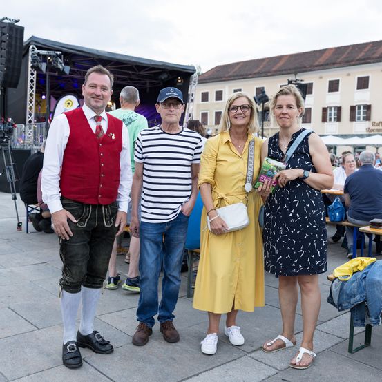Vier Menschen stehen vor einer Bühne mit Hintergrund und lächeln. Sie sind in Freizeitkleidung gekleidet. Dahinter sitzen mehrere Menschen an Tischen.