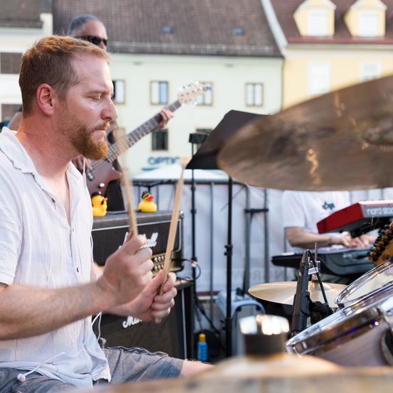 Ein Mann spielt die Drums auf einer Bühne. Ein Gitarrist und ein Keyboarder spielen hinter ihm. Gummienten sind auf dem Verstärker platziert.