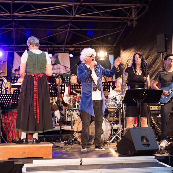 Eine Band tritt auf der Bühne auf. Der Mann in der Mitte singt in ein Mikrofon. Die Frau auf der linken Seite schaut ihn an. Hinter ihnen steht ein Orchester mit verschiedenen Instrumenten.