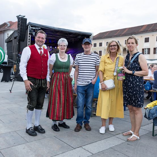 Fünf Menschen stehen lächelnd auf einem Betonplatz. Sie sind in verschiedene Outfits gekleidet. Dahinter befindet sich eine Bühne mit Lautsprechern und ein Gebäude.