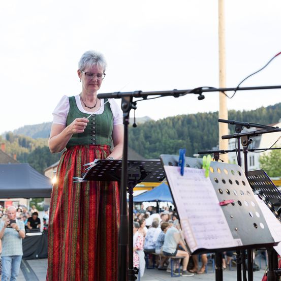 Eine ältere Frau steht auf der Bühne und hält einen Taktstock in der Hand, wahrscheinlich leitet sie ein Musikstück. Sie trägt ein traditionelles grünes und rotes Kleid. Hinter ihr sitzt ein Publikum auf Bänken und Tischen. Im Hintergrund sind Berge und Bäume zu sehen.