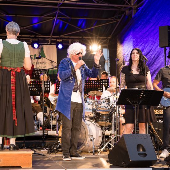 Eine Band spielt auf der Bühne, ein Mann singt in ein Mikrofon und zwei Frauen stehen daneben. Dahinter sind ein Schlagzeug und ein Gitarrist zu sehen. Die Bühne ist mit Lichtern beleuchtet und im Hintergrund befindet sich ein großes Banner.