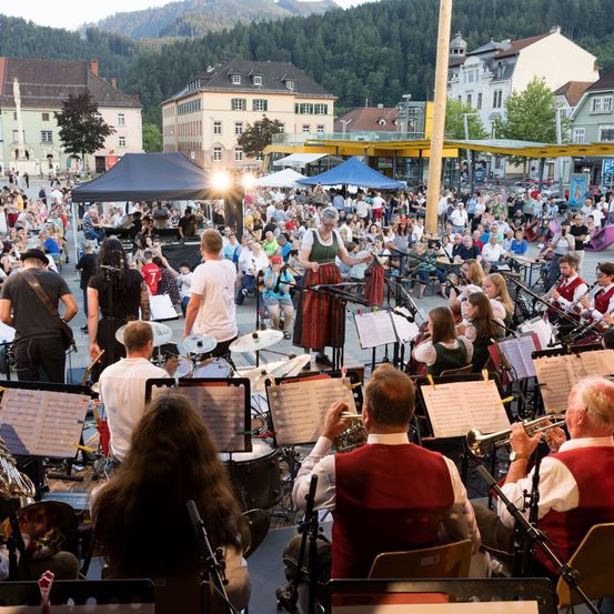 Ein großes Orchester spielt auf der Bühne in einer Freiluftveranstaltung, mit Musikern, die verschiedene Instrumente spielen. Das Publikum ist vor der Bühne versammelt, einige sitzen und andere stehen. Im Hintergrund sind Gebäude und Bäume zu sehen, und Berge sind in der Ferne sichtbar.
