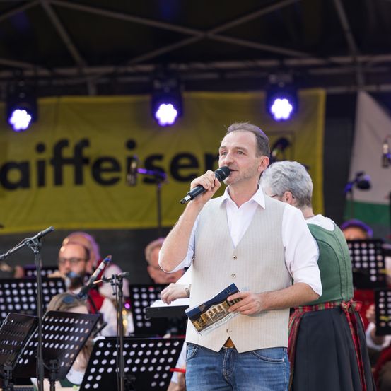 Ein Mann singt in ein Mikrofon auf der Bühne, hält ein Büchlein in der Hand, mit einer Band hinter ihm. Ein gelbes Banner mit dem Wort 'Aiffieiser' ist im Hintergrund.