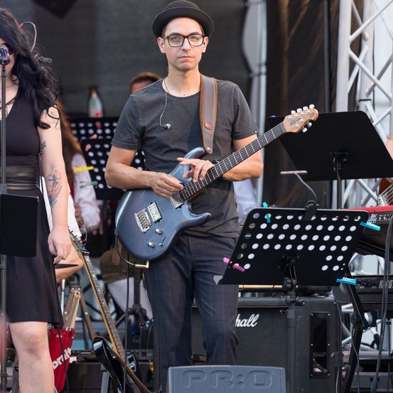 Ein Mann und eine Frau spielen Gitarre und singen auf der Bühne. Der Mann trägt einen Hut und eine Brille und hält eine blaue Gitarre. Die Frau steht links, und andere Musiker spielen dahinter.