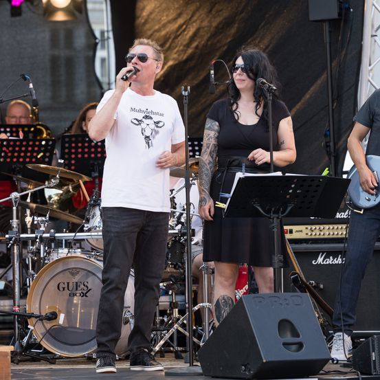 Eine Band tritt auf der Bühne auf. Der Leadsänger in einem weißen T-Shirt singt in ein Mikrofon. Eine Frau in Schwarz steht neben ihm mit einer Gitarre. Dahinter spielen andere Bandmitglieder Instrumente.