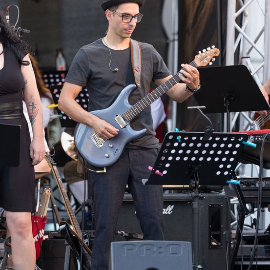 Ein Mann mit Hut spielt eine blaue E-Gitarre auf einer Bühne, während eine andere Person in der Nähe steht. Hinter ihnen sind verschiedene Musikinstrumente aufgestellt.