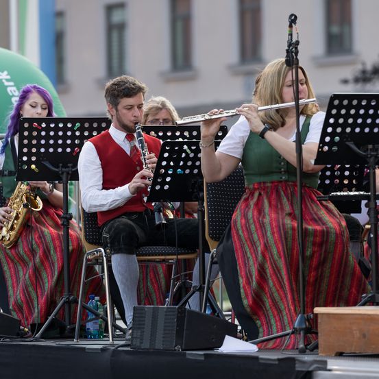 Eine Band spielt auf der Bühne, ein Mann auf einem Stuhl spielt Klarinette und eine Frau Flöte. Dahinter spielen zwei Frauen Saxophon und Trompete.