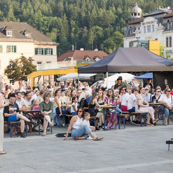Eine Menschenmenge versammelt sich im Freien unter Zelten. Viele sitzen an Tischen, einige mit Getränken, während andere stehen. Gebäude und Bäume sind im Hintergrund.