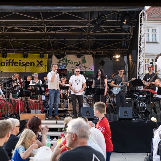 Eine Band tritt auf einer Bühne mit einem Hintergrundbild mit einem Waldlogo auf. Das Publikum sitzt davor, einige halten Getränke. Eine Frau spielt Gitarre und ein Mann singt in der Mitte.