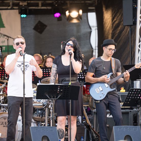 Eine Band tritt auf der Bühne auf mit einem männlichen Sänger, einer Sängerin und einem Gitarristen. Dahinter sind Trommeln, eine Trompete und ein Banner. Das Publikum ist verschwommen.