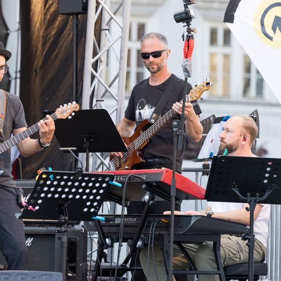 Drei Männer spielen Musikinstrumente auf einer Bühne. Einer spielt Gitarre, ein anderer spielt Keyboard und der dritte sitzt vorne.