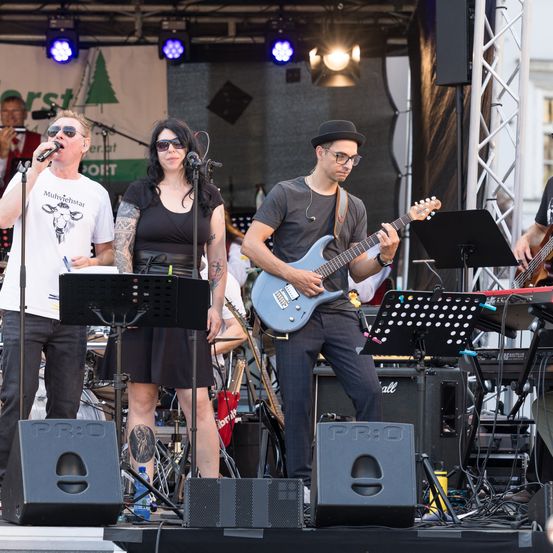 Eine Band tritt auf der Bühne auf, bestehend aus einem Gitarristen, einem Sänger und einem Keyboarder. Die Bühne ist mit Lautsprechern und Notenständern ausgestattet, mit Scheinwerfern darüber.