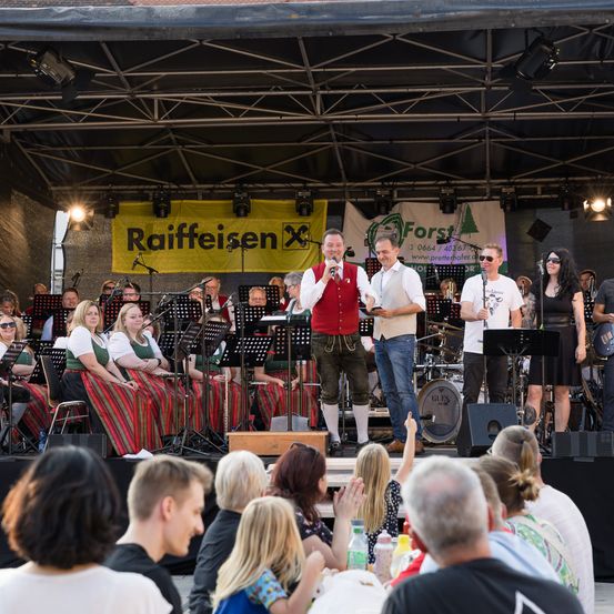 Eine Outdoor-Veranstaltung mit einer Bühnenband, zwei Männern vor Mikrofon und anderen, die Instrumente spielen. Hinter den Darstellern beobachtet eine Menge. Die Bühne hat Banner und Beleuchtung.