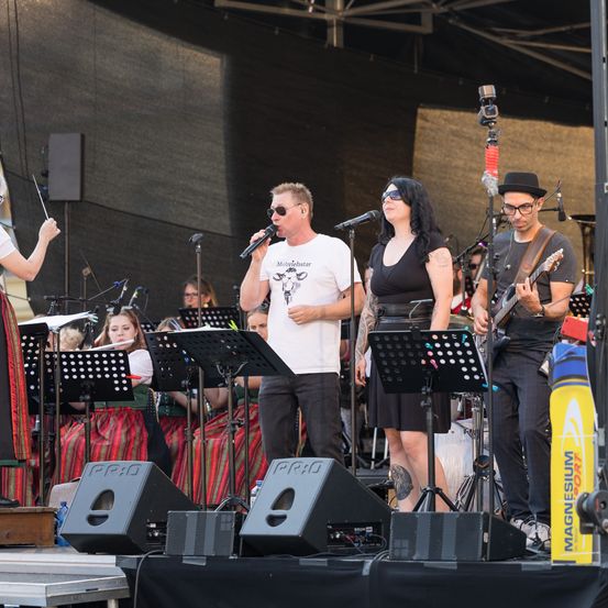 Eine Band spielt auf der Bühne mit Mikrofon und Gitarre, begleitet von einem Orchester. Die Bühne ist mit Lautsprechern und Notenständern ausgestattet, ein Dirigent führt von links.