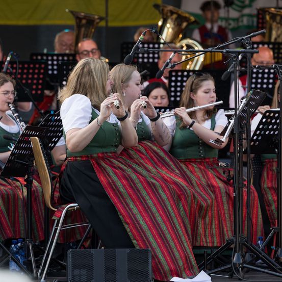 Ein Orchester mit Mitgliedern in traditioneller Kleidung spielt Instrumente. Frauen spielen Flöten. Mikrofone und Instrumente stehen auf Ständern.
