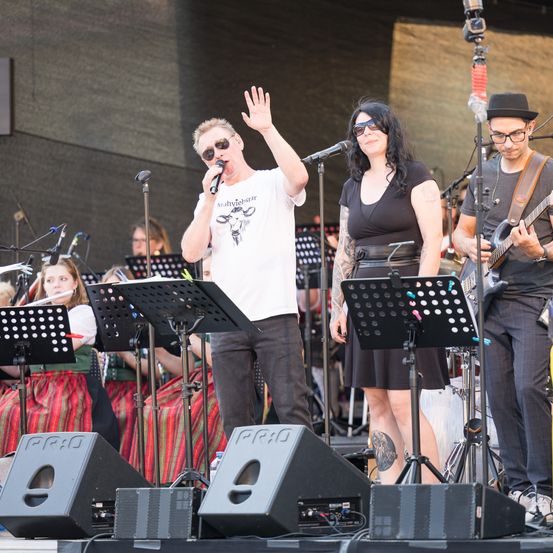 Eine Band tritt auf der Bühne auf, ein Mann singt in ein Mikrofon, eine Frau steht neben ihm, und ein Gitarrist spielt. Mehrere Musiker stehen hinter ihnen, und das Publikum schaut zu.