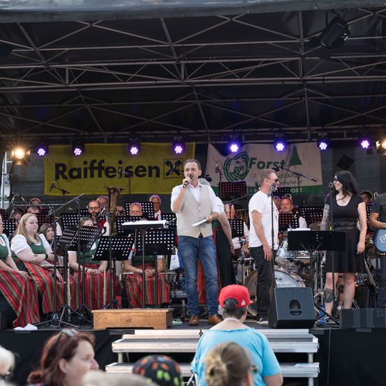 Eine Band spielt auf einer Bühne bei einer Veranstaltung im Freien. Ein Mann singt in ein Mikrofon, mit Musikern und einem Banner hinter ihm. Das Publikum sitzt auf Treppenstufen darunter.