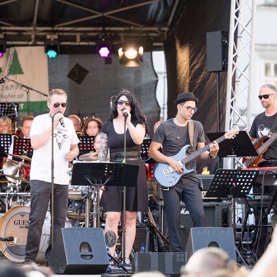 Eine Band tritt auf einer Bühne auf, mit zwei Sängern, einem Mann und einer Frau, unter bunten Lichtern. Ein Mann spielt die Gitarre und ein anderer das Keyboard. Dahinter spielt ein Schlagzeuger die Trommeln.