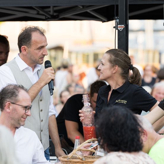 Ein Mann mit Mikrofon spricht zu einer Frau mit einer Getränkeflasche bei einer Outdoor-Veranstaltung. Menschen sitzen um einen Tisch unter einem Zelt.