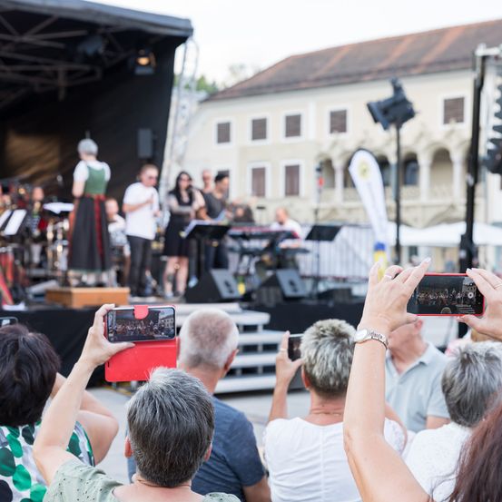 Eine Gruppe von Menschen schaut sich eine Aufführung auf der Bühne an. Einige halten ihre Telefone, um die Veranstaltung aufzuzeichnen. Im Vordergrund halten zwei Menschen ihre Telefone, um die Szene festzuhalten.