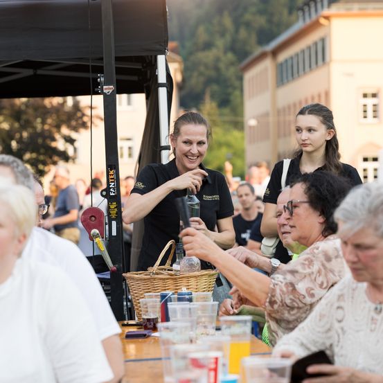 Eine Frau serviert Getränke bei einer Outdoor-Veranstaltung. Mehrere Personen sitzen um einen Tisch mit Getränken, andere sind im Hintergrund. Ein Zelt ist sichtbar.