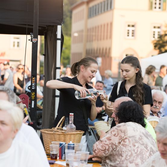 Eine Frau gießt Wasser aus einer Flasche in einen Becher auf einem Festival, mit einer anderen Frau und einer Menge im Hintergrund.