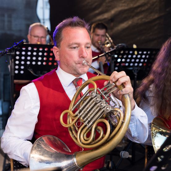 Ein Mann in einem roten Westchen spielt während eines Konzerts ein Waldhorn und schaut nach vorne. Andere Musiker im Hintergrund spielen ihre Instrumente.