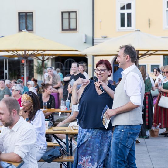 Eine Gruppe von Menschen sitzt an Tischen im Freien bei einer Veranstaltung. Eine ältere Frau spricht in ein Mikrofon, während ein Mann neben ihr lächelt. Dahinter sitzen und stehen mehrere Personen, einige mit Getränken in der Hand.