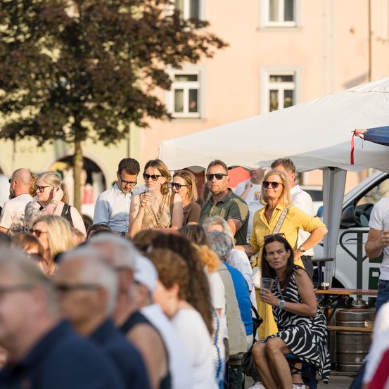 Eine Gruppe von Menschen, sowohl Männer als auch Frauen, versammelt sich im Freien, einige stehen und andere sitzen. Einige tragen Brillen, und manche halten Gläser. Ein weißes Zelt steht hinter ihnen, und einige Leute stehen in der Nähe des Zelts.
