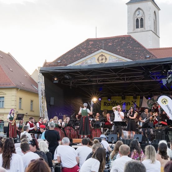 Eine Band spielt auf der Bühne, eine Menschenmenge schaut zu, unter einem Banner mit Raiffeisen, vor einem Gebäude mit Glockenturm.