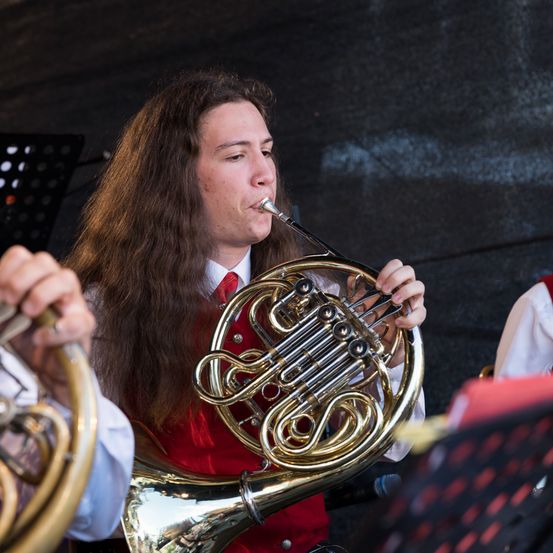 Ein Mann in einem roten Vest spielt ein Waldhorn während einer Aufführung im Freien. Eine andere Person spielt in der Nähe ein weiteres Horn.