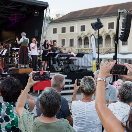 Ein Open-Air-Konzert mit einer Band auf der Bühne, Musikern, die Instrumente spielen, und Leuten im Publikum, die Fotos machen.