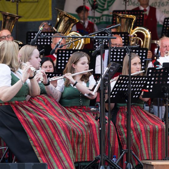 Vier Frauen in traditionellen grünen und roten Kleidern spielen Flöten auf der Bühne. Dahinter spielt eine Brassband in roten Westen und Hüten Tubas. Ein gelbes Banner hängt im Hintergrund.