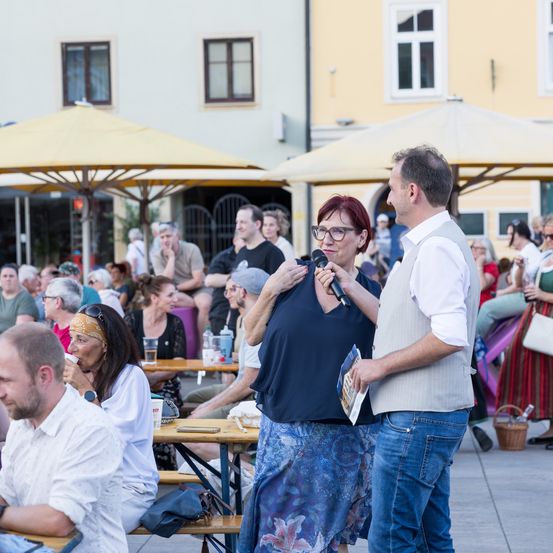 Eine Outdoor-Veranstaltung mit einem großen Publikum. Eine Frau mit Brille spricht in ein Mikrofon, während ein Mann ein Buch in der Hand hat. Im Hintergrund sitzen und stehen verschiedene Menschen.